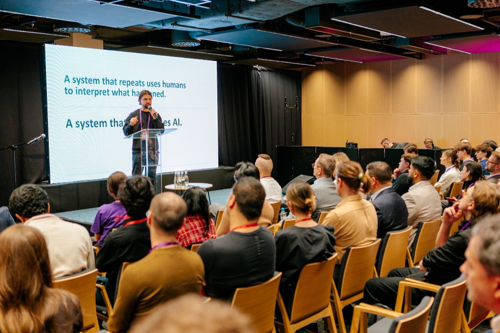 Macklin Buckler speaking on stage at MIT Universal AI conference in Warsaw, approximately 60 attendees visible, slide reads: A system that repeats uses humans to interpret what happened. A system that learns uses AI.